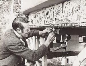 Black and white photograph of a person fixing and decorating the outside of their old-style caravan.