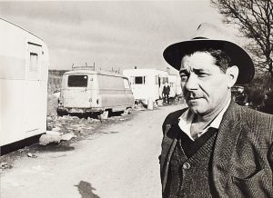 Black and white photograph of person in the foreground in front of caravans parked on road.