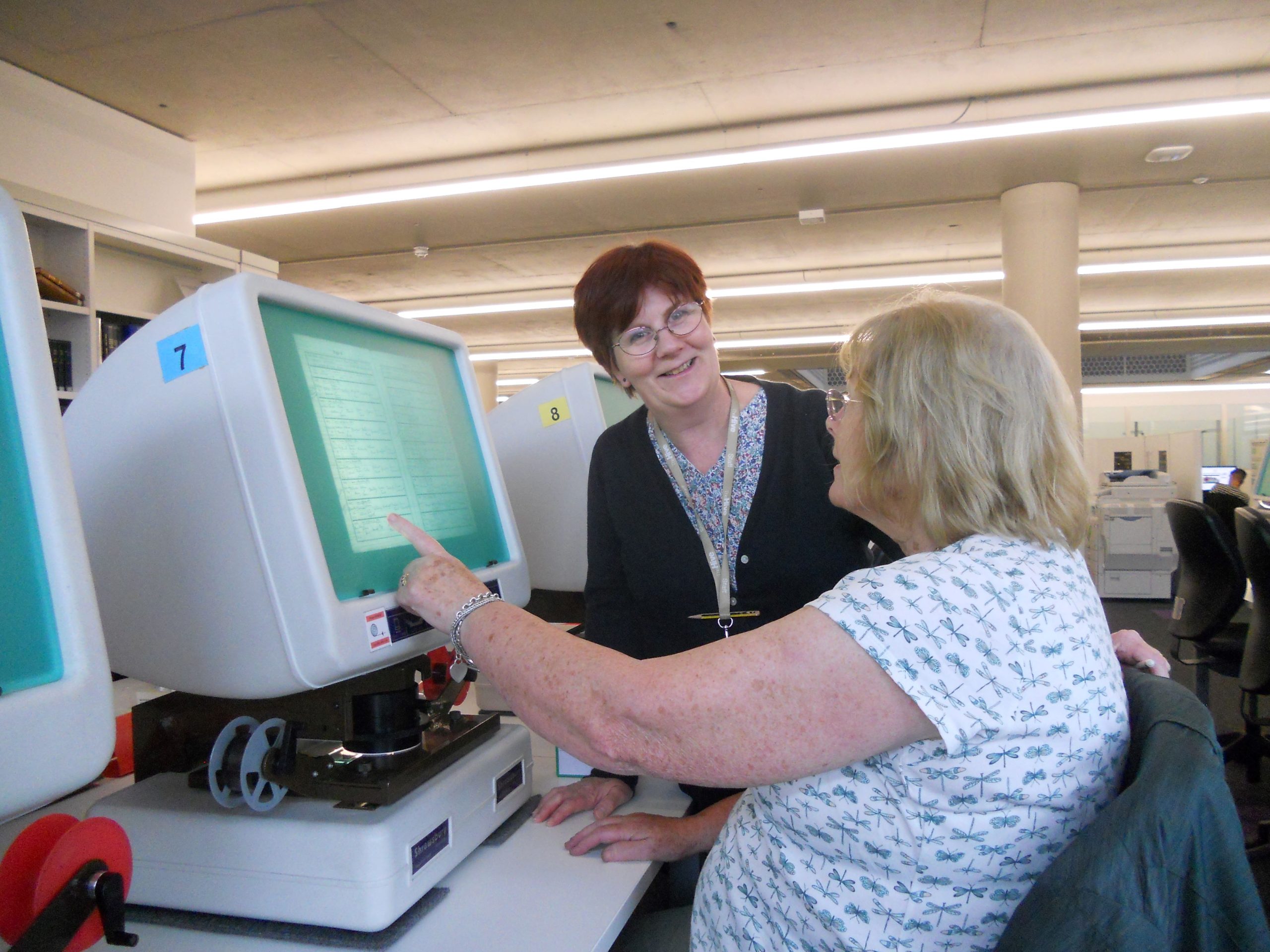 Colour photograph showing a member of staff helping a customer consult a microfilm on a reader.
