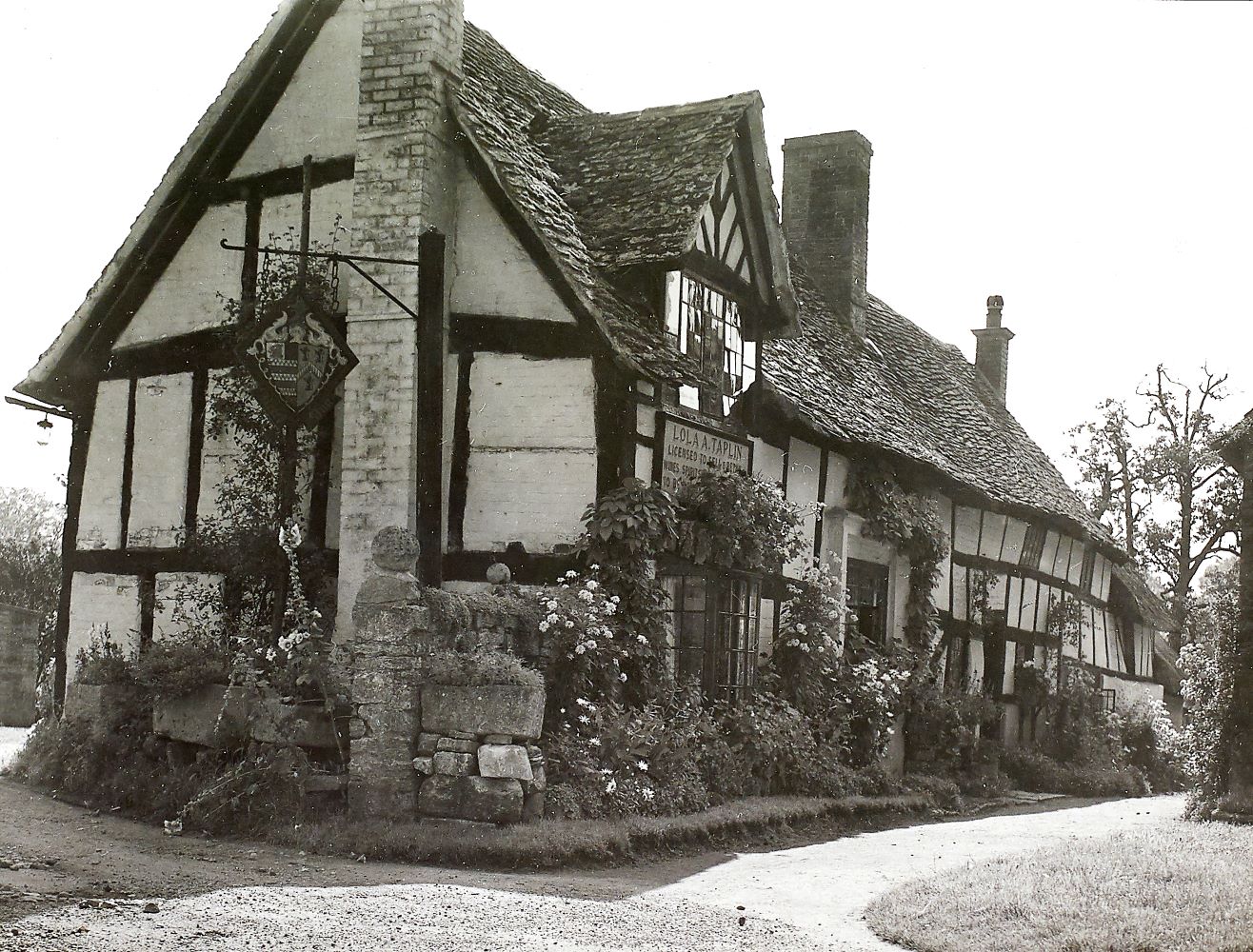 Black and white photo of an old timber framed building with a wobbly tiled roof and plants outside