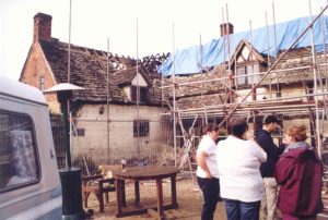 Pub with scaffolding around it, blue tarpaulins on the roof and a small ground of people standing outside (righthand side of image) 