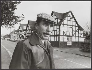 Black and white photo of the top half of a man with a moustache wearing a flat cap standing on a street with houses on